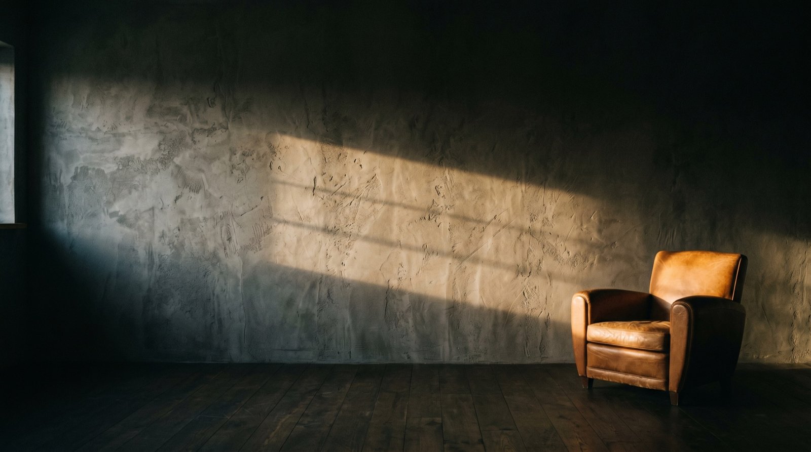 A hand trowelled microcement wall in raking light, a leather armchair on dark timber boards in front of it